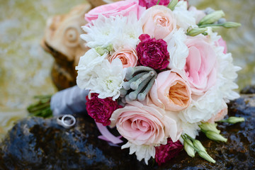 Close up of bridal bouquet of pink, red and white roses and wedding rings on stone background outdoors, copy space. Wedding concept