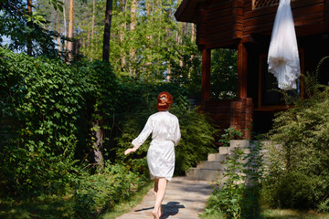 Fototapeta na wymiar Beautiful redhead bride in silk dressing gown running barefoot to her wedding dress hanging on wood house, copy space. Wedding morning preparation in sunny summer day