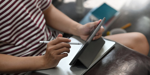 Cropped image of creative man drawing on computer tablet by stylus pen while sitting at the leather couch with quiet sitting room as background.