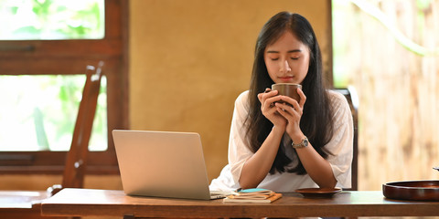 Woman holding a coffee cup while sitting in front her computer laptop that putting on wooden working desk and surrounded by stack of notebook and pen.