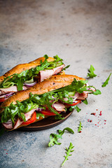 Two fresh baguette sandwiches with meat, tomato, cucumber and arugula on gray background.