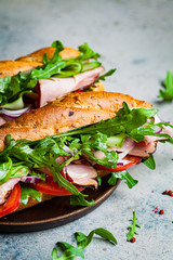 Two fresh baguette sandwiches with meat, tomato, cucumber and arugula on gray background.