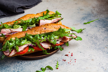 Two baguette sandwiches with meat, tomato, cucumber and arugula on gray background.