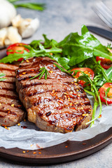 Grilled beef steaks with arugula and tomato salad on wooden plate, dark background.