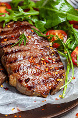 Grilled beef steaks with arugula and tomato salad on wooden plate, dark background.