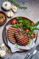 Grilled beef steaks with arugula and tomato salad on wooden plate, dark background, top view.