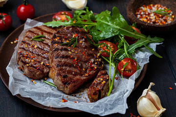 Grilled beef steaks with arugula and tomato salad on wooden plate, dark background.