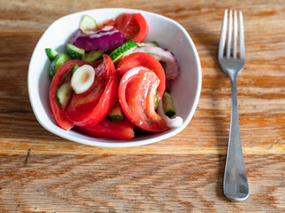 top view of portion of homemade salad from fresh vegetables (sliced and oiled tomato, cucumber, onion) in white bowl on shabby wooden table at home kitchen