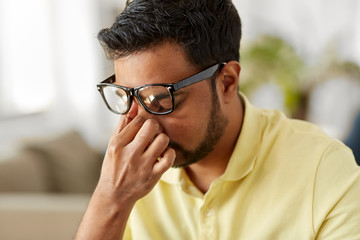 fatigue and people concept - tired young indian man in glasses rubbing his nose bridge at home