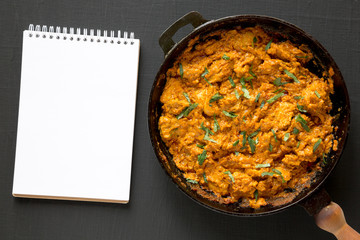 Homemade Chicken Tikka Masala in a cast iron pan, blank notepad on a black surface, top view. Flat lay, overhead, from above.