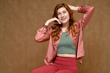 Fototapeta premium Nice girl with long hair dressed in red trousers and a pink blouse. Portrait of a young Caucasian red-haired woman sitting on a stand on a beige background in various poses.