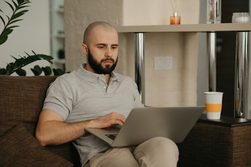 Fototapeta premium Remote work. A man working remotely on his laptop. A bald businessman with a beard works sitting on a sofa at home.