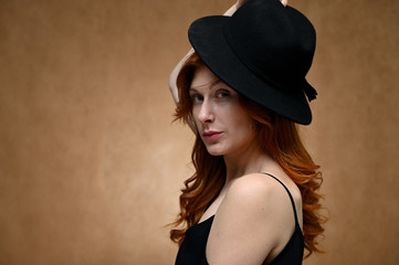 Studio large photo portrait of a caucasian woman with long red hair on a beige background. Model posing in a hat.