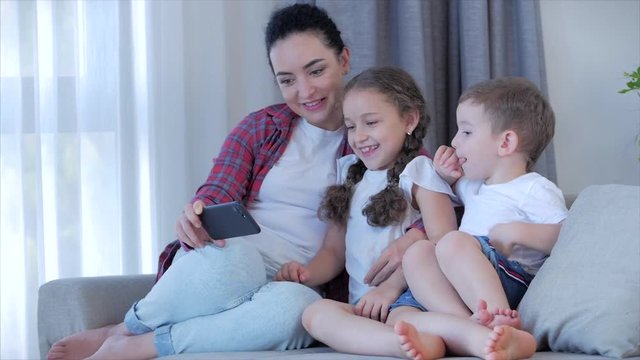 Happy family,mom and cute little kids,mother playing with children at home relaxing use a smartphone cuddling sit on sofa daughter and son laugh,watch funny videos,have fun,enjoy family life moments