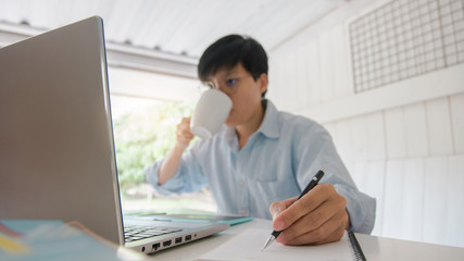Asian young businessman of a student holding a pen writing letter on notebook and drinking hot coffee, he reading news globally on the digital laptop in the morning at home.