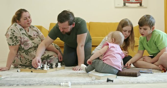 A large family with many children spends time together during self-isolation in quarantine. Children play chess and look at photos by the sofa.