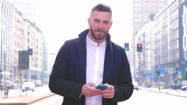 Slow motion of young handsome caucasian bearded man outdoor holding smartphone looking camera smiling – 5g technology, confident, positive concept