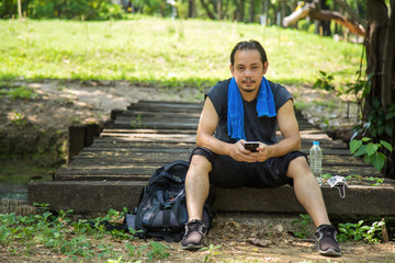 Obraz premium An Asian man in a black sportwear with blue towel on his neck sitting on wooden branch bridge with backpack and face mask, water bottle at side in public park.