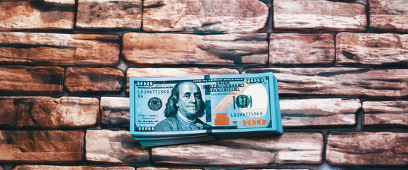 A pack of dollars.A large wad of hundred-dollar bills is lying on a brick wall of dark red brown color.