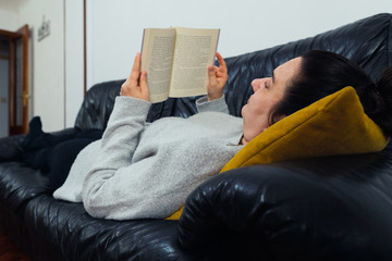 Middle-aged white woman reading a book lying on a black sofa with a yellow cushion. She is wearing a gray sweater.
