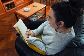 Middle-aged white woman reading a book lying on a black sofa with a yellow cushion in her spacious living room. She is wearing a gray sweater.
