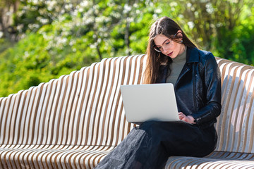 young elegant woman in eyeglasses sitting on bench in park and working on laptop on a sunny and windy day