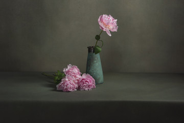 Vintage pottery vase with one peony and some peonies lying next to it on a table in a grey room.