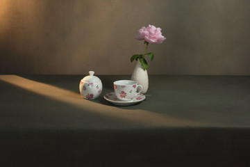 Tea cup with saucer, sugar bowl and white vase with peony on a table in a grey room in morning sunlight.