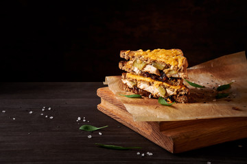 Chicken and caramelized onion sandwich with melted cheese with some greens and sea salt, on piece of baking paper on wide wooden chopping board on dark brown table with dark background