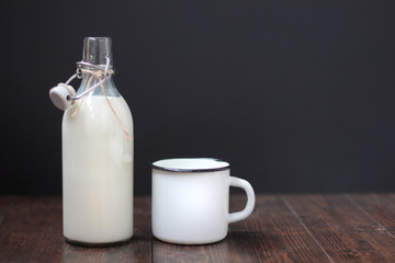 Glass bottle of milk and white metal mug on a dark rustic wooden table. Black background
