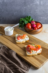 Sandwiches from bread toasts with cereals and bran, cottage cheese and red fish. Vertical still life of two slices of delicious snacks on a wooden cutting board, bowl of vegetables on a marble table.
