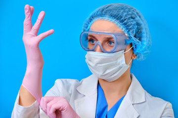 A nurse on a blue background puts on medical gloves. Doctor in a protective mask and clothing for the treatment of coronavirus, close-up portrait