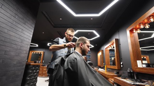 Trendy barber cuts bearded man's hair with a clipper in barbershop. Men's hairstyling and hair cutting in salon. Grooming the hair with trimmer. Hairdresser doing haircut in retro hair salon. Tracking