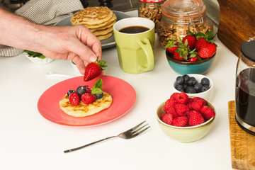 Mano de hombre sosteniendo una fresa, preparando el desayuno con tortitas junto a una taza de café, nueces y frutos rojos en la cocina. Vista superior