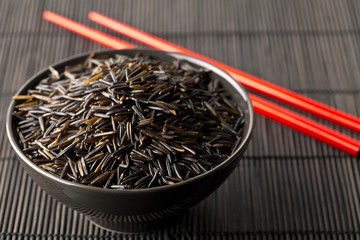 Heap of uncooked, raw, black wild rice grains in black bowl with red chopsticks on black bamboo mat