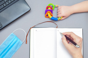 Fototapeta na wymiar Top view of hand of man writing to notebook while hand of child holding rattle toy with protective medical mask and laptop on gray background. Work from home.