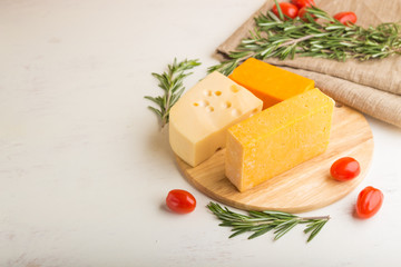 Various types of cheese with rosemary and tomatoes on wooden board on a white wooden background. Side view, copy space.