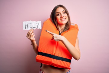 Young beautiful brunette woman wearing lifejacket holding paper with help word message very happy...