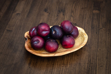 Beautiful ripe plums in a wooden plate on a table top made of boards with a dark textured surface.