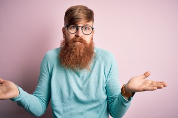 Handsome Irish redhead man with beard wearing glasses over pink isolated background clueless and...