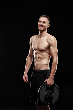 Strong athletic smiling man - athlete fitness model showing his perfect body isolated on black background with copyspace. Ectomorph bodybuilder holding a black barbell in his hands.