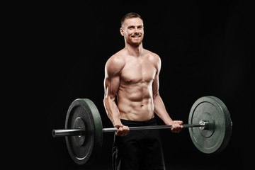 Fototapeta na wymiar Strong smiling athletic man - crossfit athlete fitness model showing his perfect body isolated on black background with copyspace. Shakes biceps, holds barbell in hands, perfect shoulders and chest