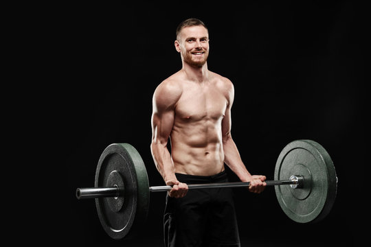 Strong smiling athletic man - crossfit athlete fitness model showing his perfect body isolated on black background with copyspace. Shakes biceps, holds barbell in hands, perfect shoulders and chest