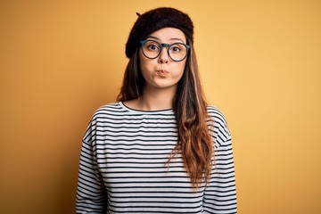 Young beautiful brunette woman wearing french beret and glasses over yellow background puffing cheeks with funny face. Mouth inflated with air, crazy expression.