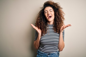 Young beautiful woman with curly hair and piercing wearing casual striped t-shirt celebrating mad...