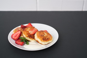 Russian and Ukrainian cuisine. Breakfast. Cottage cheese pancakes. Cheesecakes with fresh strawberries, mint and sour cream sauce in a white plate on a light grey background. Copy space mocap close-up