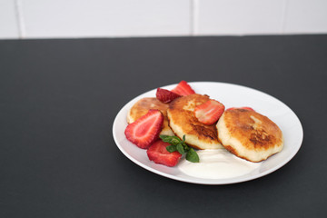 Russian and Ukrainian cuisine. Breakfast. Cottage cheese pancakes. Cheesecakes with fresh strawberries, mint and sour cream sauce in a white plate on a light grey background. Copy space mocap close-up