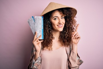 Young beautiful woman with curly hair and piercing wearing asian hat holding boarding pass surprised with an idea or question pointing finger with happy face, number one
