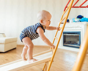 Cute baby performs gymnastic exercises on a wooden home sports complex stairs and rings. Children's sports exercises. Physical education of children at home