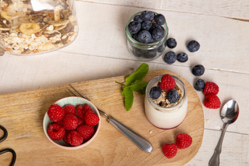 White yogurt glass jar with fresh raspberries and blueberries on serving board on rustic table, wooden tray as background, copy space